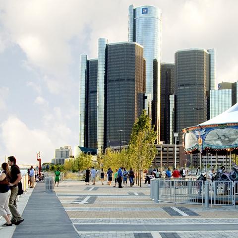 Side-view of the Detroit RiverWalk, including the river, pedestrians, a carousel, and office buildings in the background.